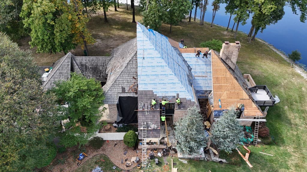 Chad Schwarz residence — aerial documentation mid-restoration: cedar shake tear-off, bright synthetic underlayment, new charcoal shingles in progress, crew on roof